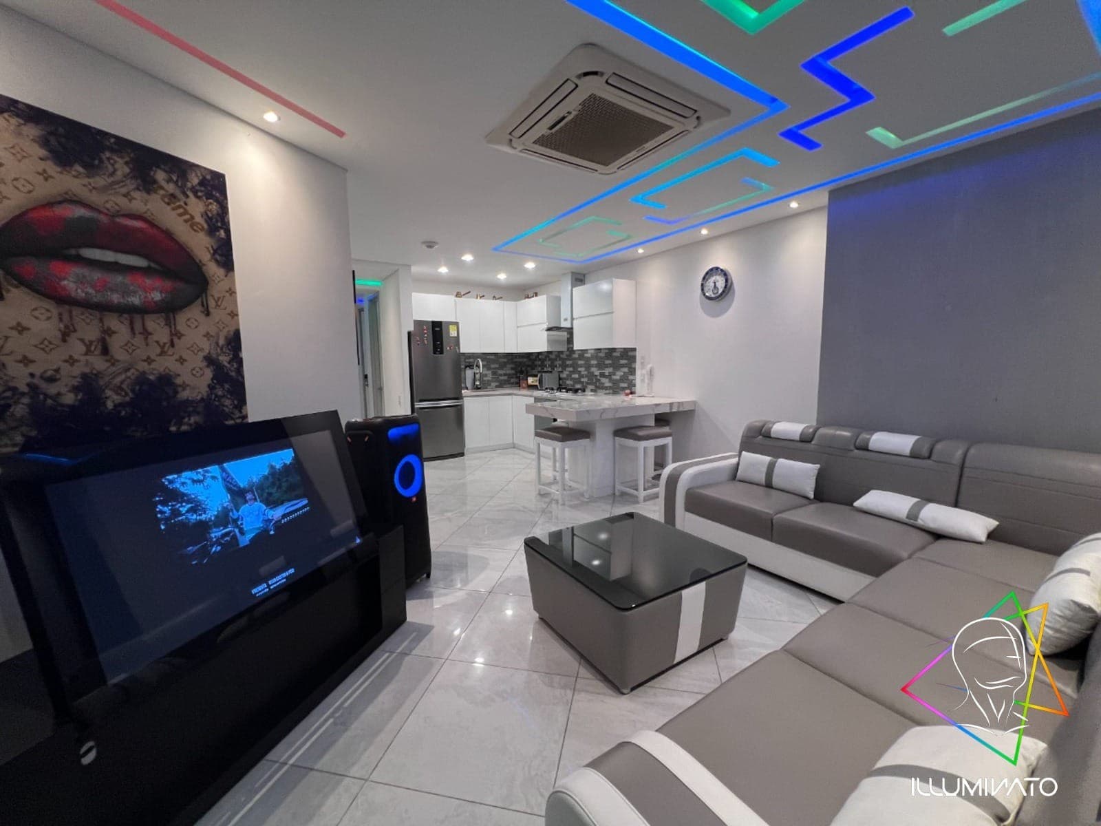 Sala con sofá modular en L, televisor grande y la cocina integral al fondo.