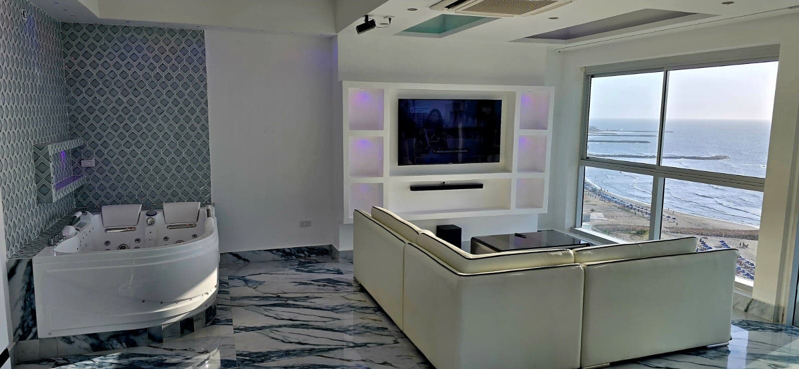 Sala con sofá blanco en L, televisor empotrado y jacuzzi junto al ventanal.