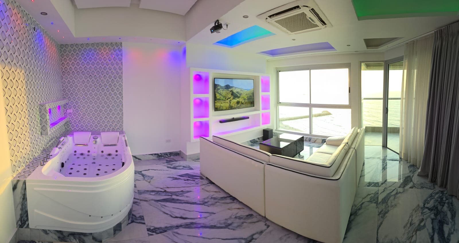 Zona social con jacuzzi, televisor y vista al mar desde el ventanal.
