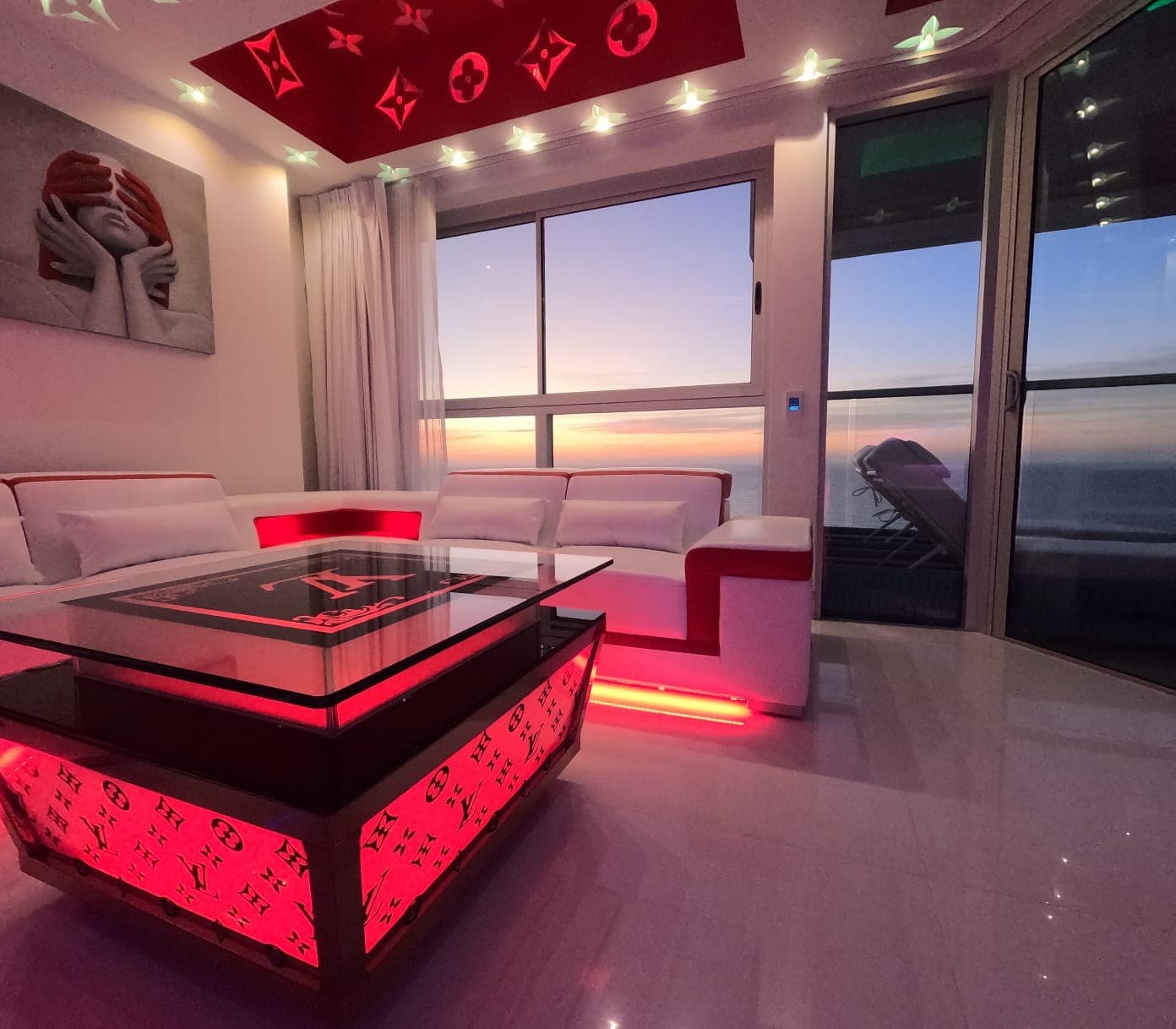 Sala con sofá blanco, mesa de centro iluminada en rojo y ventanal al mar.