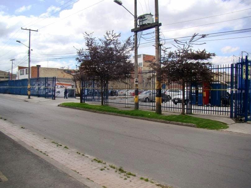Calle frente al lote, con el cierre en reja y árboles en el andén.