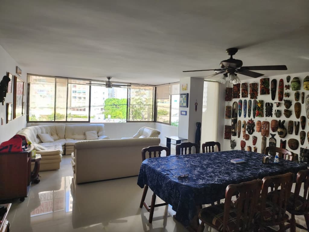 Sala-comedor con ventanales, ventilador de techo y una colección de máscaras en la pared.